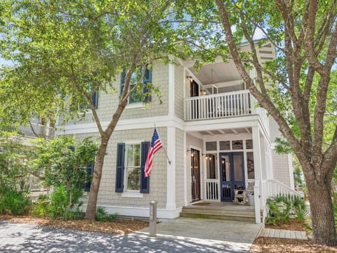 Charming two-story coastal home with classic shingle siding, welcoming front porch, and mature shade trees creating a peaceful neighborhood setting.
