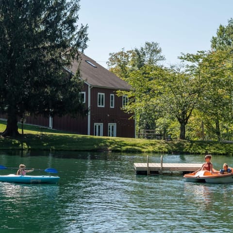 Private 1/3-acre fishing pond behind the restored barn featuring 3 kayaks, a paddle boat, and swim dock. Life jackets included. Enjoy shaded pond views under a towering pine on 5+ private green acres, perfect for nature stays and peaceful getaways.