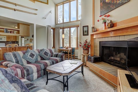 A cozy living room with patterned sofas, a wooden coffee table, a fireplace, and large windows allowing natural light. The kitchen and dining area are visible in the background.