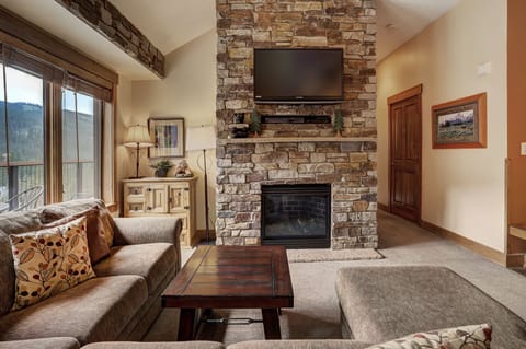 A cozy living room with a stone fireplace, mounted TV, sectional sofa, wooden coffee table, and a window with a view of the mountains.