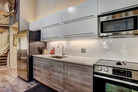 A modern kitchen featuring stainless steel appliances, white upper cabinets, wooden lower cabinets, a sink, and an electric stove with a microwave above. A black refrigerator stands to the left.