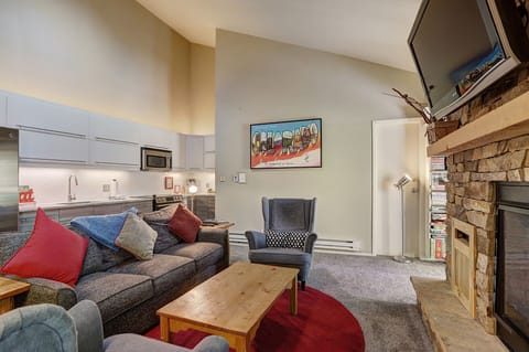 A cozy living room with a gray sofa, colorful throw pillows, a wooden coffee table, and an armchair. The kitchen is visible in the background, and there's a fireplace with a TV mounted above it.