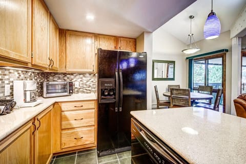 A kitchen with wooden cabinets, a black refrigerator, a microwave, and a dishwasher. A dining area with a table and chairs is visible in the background, with blue pendant lights above the counter.