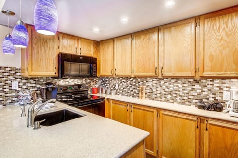 A modern kitchen with wooden cabinets, a mosaic tile backsplash, black appliances, a beige countertop with a sink, and blue pendant lights.