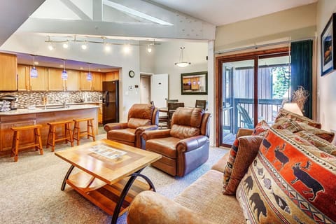 Spacious living room with two leather recliners, a patterned sofa, coffee table, and view of kitchen area with wooden bar stools and blue pendant lights. A dining table is near a sliding glass door.