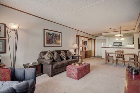 A living room with a brown sofa, patterned cushions, floor lamp, artwork on the wall, and a wooden dining table in the background near a kitchen area.