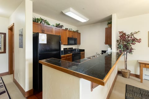 A kitchen with dark countertops, brown cabinets, a black refrigerator, and a black stove. A small indoor plant is placed in the corner.