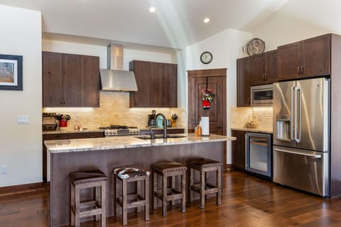 A modern kitchen with dark wood cabinets, stainless steel appliances, a marble island with three stools, and hardwood floors. A wreath decorates the door, and a clock is visible on the wall.