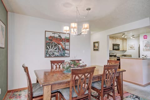 A dining room with a wooden table and six chairs, an overhead light fixture, wall art featuring a red tractor, and a view of a kitchen with light-colored cabinetry and appliances.