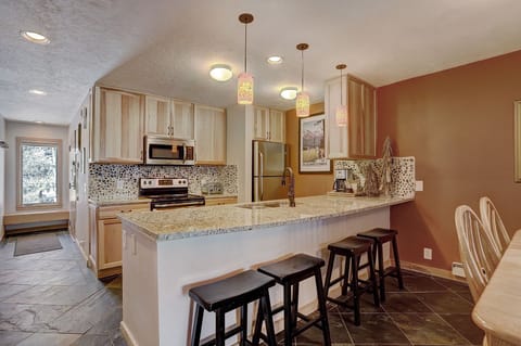 A modern kitchen with light wood cabinets, stainless steel appliances, a mosaic tile backsplash, and a granite countertop with three black stools. Pendant lights hang above the counter.