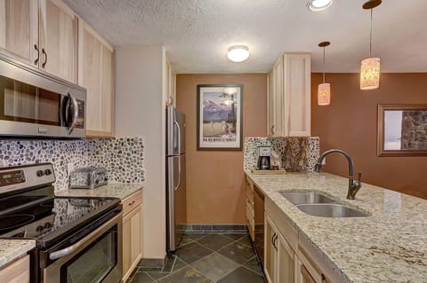 A modern kitchen with stainless steel appliances, granite countertops, stone tile backsplash, and wooden cabinets. There are pendant lights above the sink area and a framed picture on the wall.