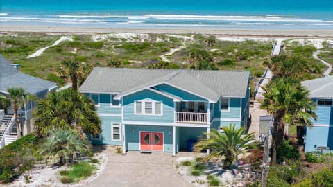 Oceanfront exterior of a blue two-story house with coral double doors, balcony seating, and sandy beach access.