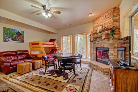 A cozy living room features a stone fireplace, a round table with chairs, a bunk bed, and a leather sofa. A ceiling fan, windows with a forest view, and a TV on a stand complete the room.