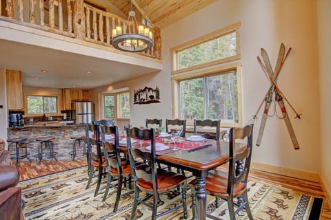 A dining room features a wooden table with eight chairs, ski decor on the wall, large windows, and an adjoining open kitchen with bar stools and stone accents. Natural light fills the space.
