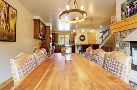 A dining room with a long wooden table, upholstered chairs, a circular chandelier, and a view of the kitchen. A stone fireplace and framed artwork are visible on the right wall.