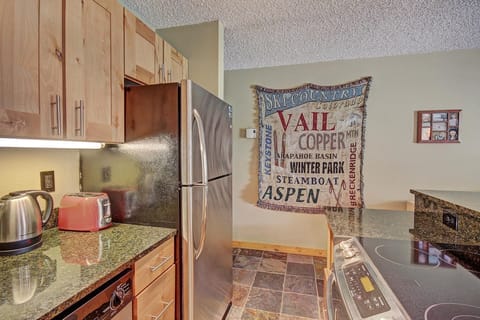 A kitchen with wooden cabinets, stainless steel fridge, stove, kettle, red toaster, and a granite countertop. A tapestry featuring ski resort names hangs on the wall next to a small photo display.