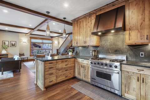 A modern kitchen with wooden cabinets and stainless steel appliances, adjacent to an open living area with wooden beams, stairs, and seating.