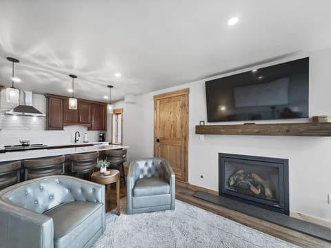 A modern living room with two gray armchairs, a wooden coffee table, a wall-mounted TV above a fireplace, and a view into the kitchen with dark cabinets and pendant lights over a countertop.