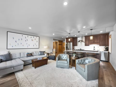 Modern living room with sectional sofa, two armchairs, a wooden coffee table, wall art, and an open-plan kitchen with wooden cabinets, stainless steel appliances, and a small dining area.