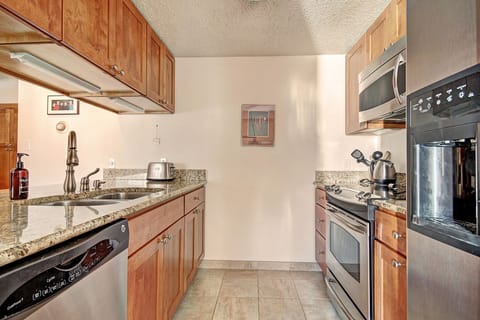 Modern kitchen with stainless steel appliances, granite countertops, wooden cabinets, and beige tile flooring. A wall-mounted microwave and oven are on the right side, and framed art decorates a wall.
