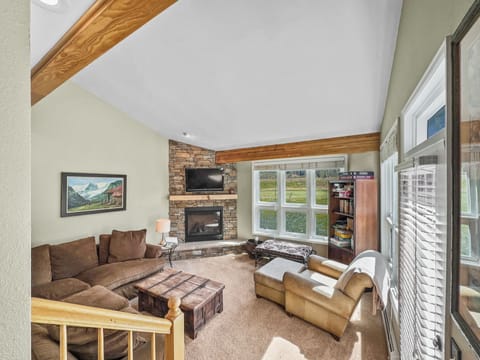 A living room with a brown sectional sofa, a stone fireplace, a wooden coffee table, and a bookshelf. The room features a large window and exposed wooden beams on a high ceiling.