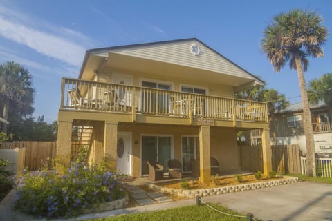 Two-story villa exterior with upper deck featuring beige railings and patio seating below on textured stucco pillars.