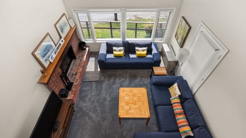 A bird's-eye view of a living room with two blue sofas, a fireplace, and a wooden coffee table. Large windows offer an outside view, and the room has framed artwork and a floor lamp.