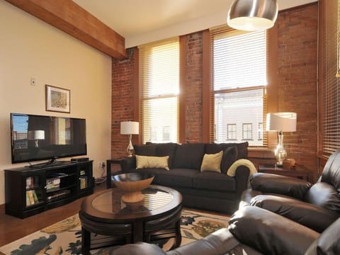 Welcome to your historic loft retreat! Relax in this cozy living room featuring beautiful exposed brick walls, towering wood-beamed ceilings, and plush leather seating.