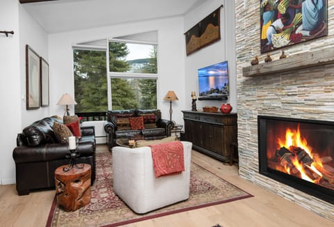 Cozy up by your crackling fireplace while enjoying forest views through expansive windows in this inviting living room.