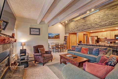 A spacious living room with a teal sectional sofa, colorful pillows, mounted TV, and coffee table. In the background, a dining area and kitchen with wooden cabinets and stainless steel appliances.