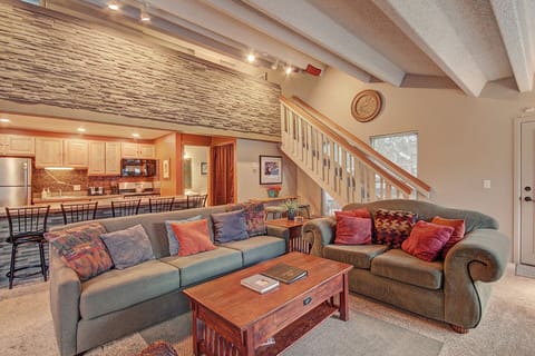 A living room with a green sofa, loveseat, and wooden coffee table. Stairs with a wooden railing lead to an upper level. The kitchen, viewable in the background, has wooden cabinets and a stone backsplash.