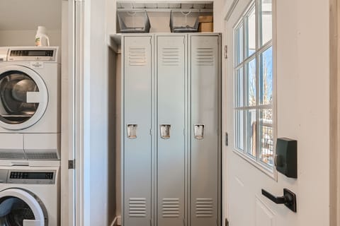 A laundry room with a stacked washer and dryer on the left, and three gray lockers next to a door with a window on the right.