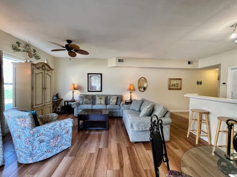 Unwind in your spacious living room with plush sectional seating, tropical ceiling fan, and warm natural light streaming through large windows.