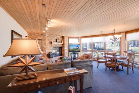 Unwind in your cozy living space with warm wood ceiling, crackling fireplace, and panoramic windows showcasing the city beyond.