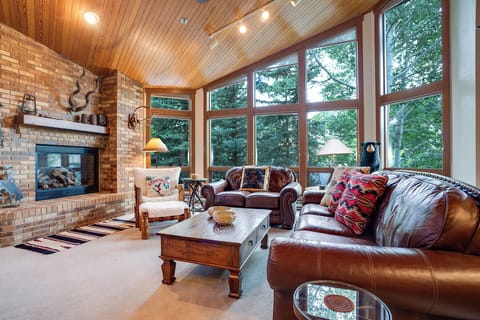 Sink into rich leather sofas beside a crackling fireplace, surrounded by soaring windows that frame lush forest views in this cozy living space.