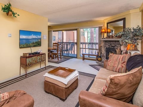 Living room with earth-toned sofa, stone fireplace, and sliding glass doors opening to a balcony with outdoor seating.