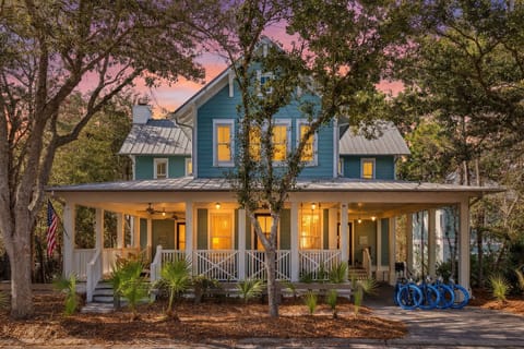 Charming blue cottage nestled among oak trees with wraparound porch and bicycles ready for coastal adventures at sunset.