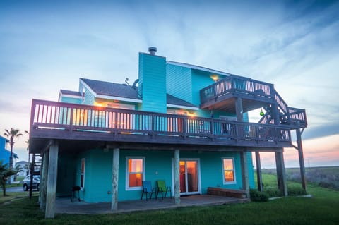 Vibrant turquoise beach house with expansive wraparound decks overlooking coastal grasslands during golden hour twilight.