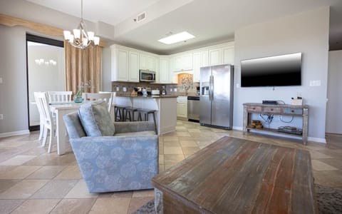 Bright living space features a comfortable blue armchair, dining table, and a modern kitchen with sleek cabinetry and a TV