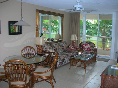Relax in your open-concept living and dining area featuring tropical rattan furnishings, ceiling fan, and glass doors framing lush palm views outside.