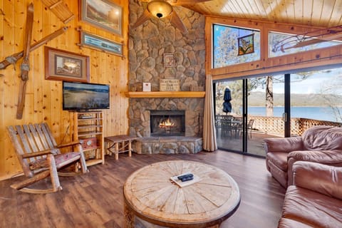 Cozy cabin living room with a stone fireplace, comfy seating, and a TV with an amazing view to the lake—perfect for relaxing evenings, movie nights, and unforgettable mountain getaways.