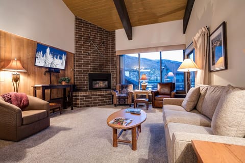 Relax in your cozy living room with exposed beams, brick fireplace, and stunning mountain views through floor-to-ceiling windows.