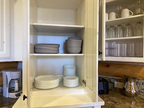 The white kitchen cabinets have a fresh, clean look, offering a modern yet cozy contrast to the rustic charm of the cabin. Neatly arranged, you’ll find a set of glass cups, mugs, plates, and bowls.