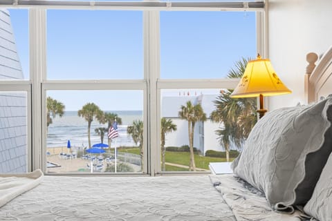 Master bedroom with white quilted bedding and a palm tree lamp, overlooking ocean and pool area.