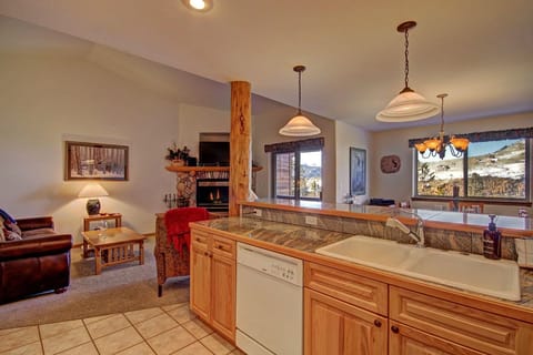 A cozy open-plan living room and kitchen area with wooden cabinetry, modern appliances, a leather sofa, a stone fireplace, large windows, and mountain views.