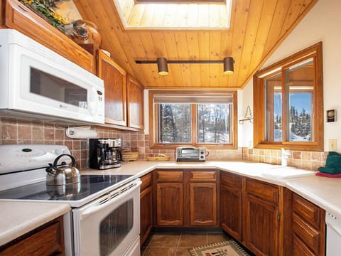 Your mountain cabin kitchen awaits with warm wood accents, modern appliances, and stunning snowy forest views through picture windows.