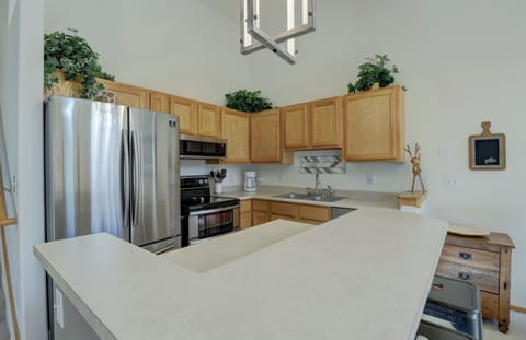 A kitchen with light wood cabinets, stainless steel appliances, a double-door refrigerator, and a light-colored countertop. Potted plants are placed above the cabinets as decoration.