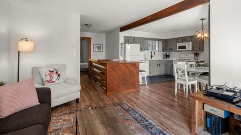 A modern kitchen and living area with wooden flooring, a mix of light and dark furniture, and a large wooden island. The kitchen is equipped with white appliances, and there's a white dining set nearby.