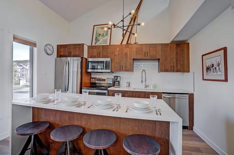 A modern kitchen with wooden cabinets, a white island with four stools, set for dining. Stainless steel appliances, a contemporary light fixture, and framed artwork are visible.