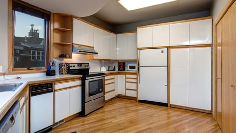 A modern kitchen with white cabinets, stainless steel appliances, a built-in oven, a refrigerator, a dishwasher, a microwave, and wooden flooring. A window offers an outside view.